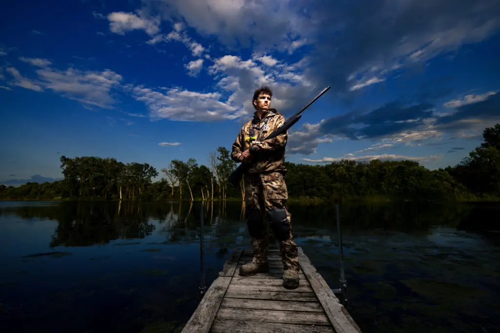 Male high school senior wearing hunting gear and posing by a lake during blue hour in Owatonna, Minnesota, for a senior portrait session.