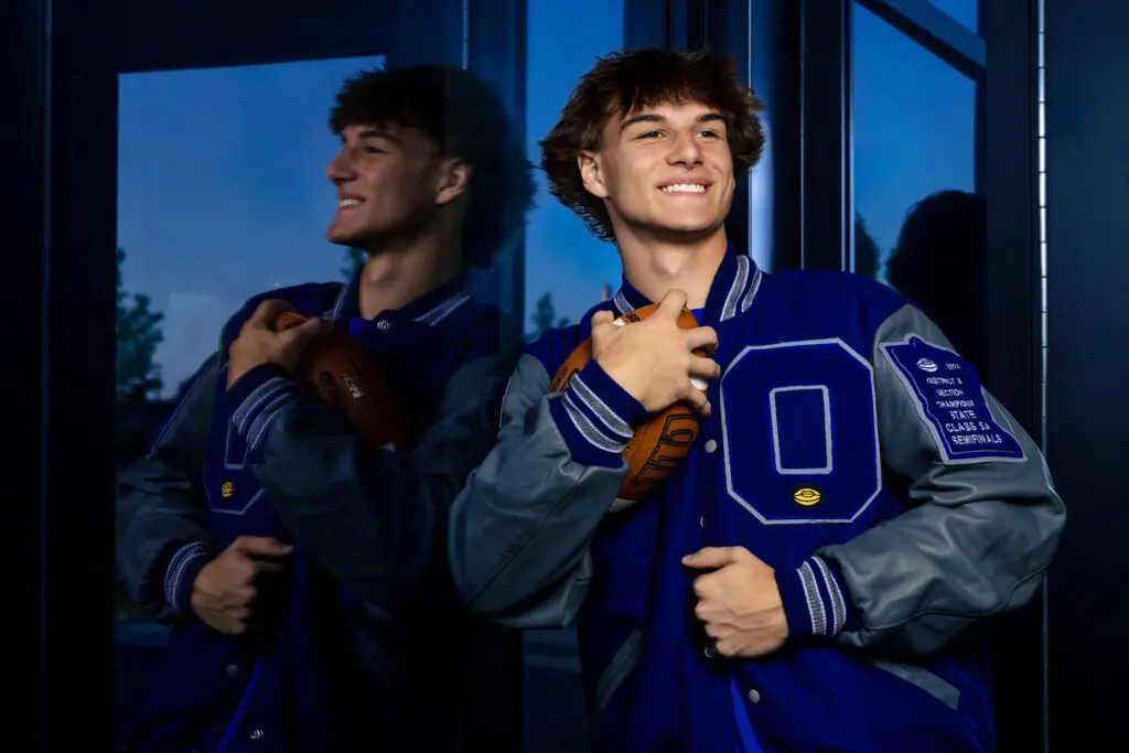Male high school senior in Owatonna, Minnesota, posing against a wall with a reflection while holding a football and wearing a letterman jacket during a senior portrait session.