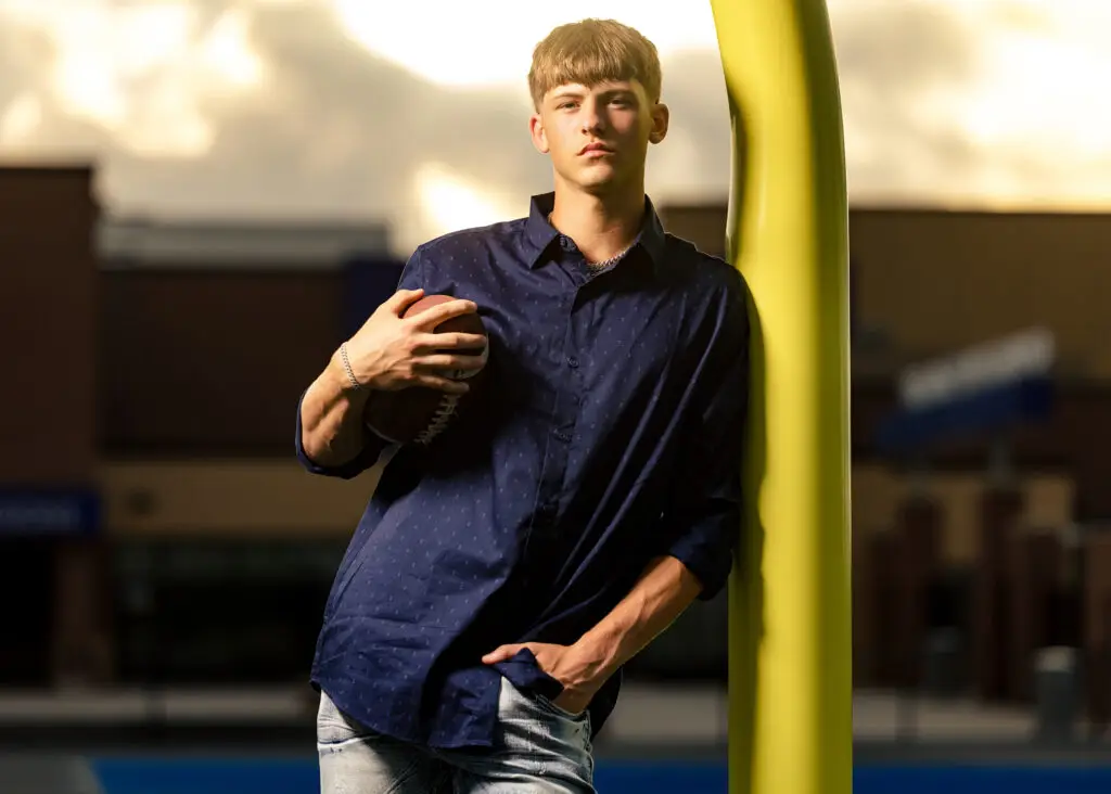 Male high school senior posing on Owatonna Federated Football Field with a football leaning against the goalpost during a senior portrait session.